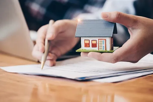 One hand holds a small model house while the other signs papers spread across a desk