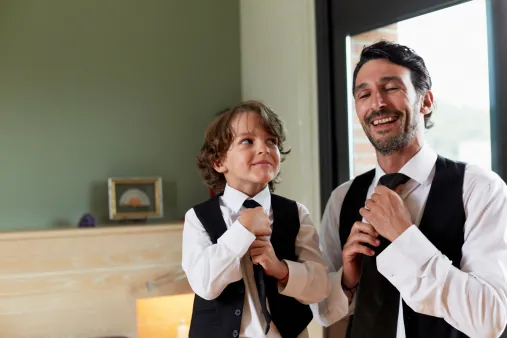 A boy in a waistcoat ties his tie beside a smiling man, copying him, both in white shirts