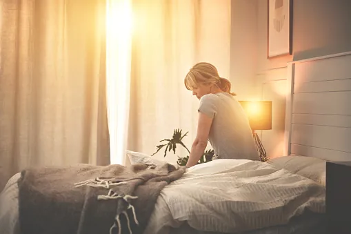 A woman sits on the edge of an unmade bed, head bowed, as morning sun streams through the curtains