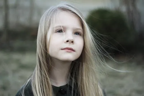 A girl with long fair hair stands outdoors, looking into the distance as the wind lifts her hair