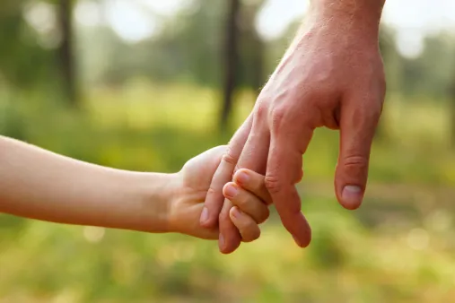 A small child's hand holding on to an adult's finger, outdoors against blurred green