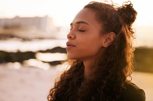A woman with curly hair stands by the water with her eyes closed, breathing calmly in the evening light