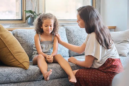 A woman on a sofa rests a hand on the shoulder of a girl who is looking down at her own hands