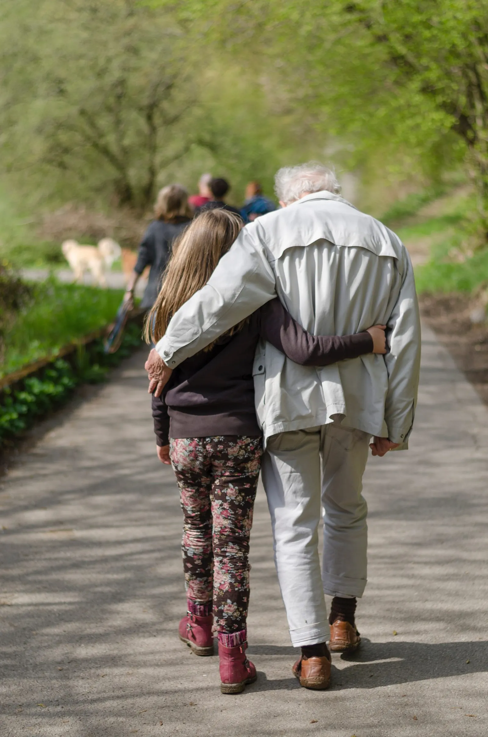 Seen from behind, a girl and a white-haired older adult walk arm in arm down a leafy gravel path