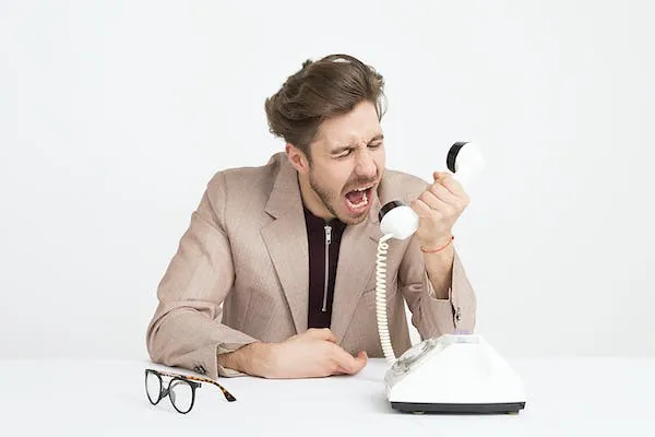 A man in a beige blazer shouts into the handset of an old white rotary phone, his glasses on the table