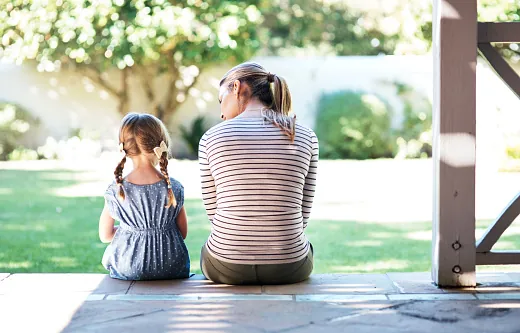 A woman and a girl with braids sit side by side on a porch step, seen from behind, facing a sunny garden