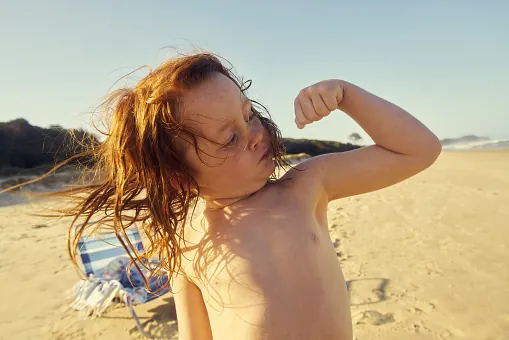 A red-haired child on a sunny beach flexes an arm and looks at their own muscle