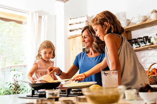 A woman cooks pancakes at the hob in a sunny kitchen while two girls help with the pan and the plate