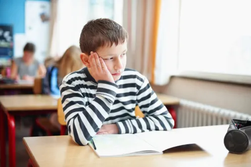 Boy at a school desk resting his cheek on his hand, looking away from the open book in front of him