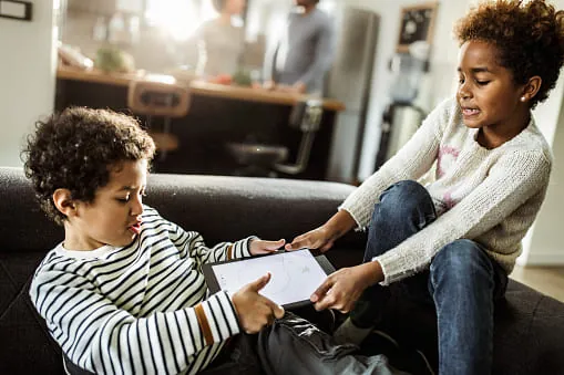 Two children on a sofa tugging at the same tablet, with adults blurred in the background
