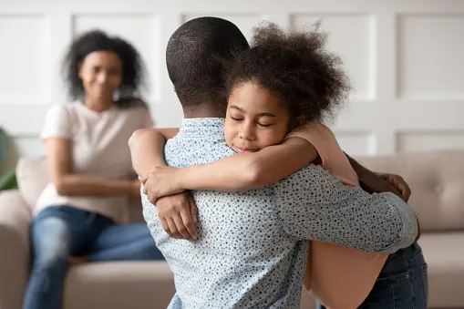 A girl hugs a man tightly with her eyes closed while a woman watches from the sofa behind them
